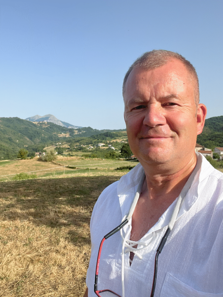 a photo of Chris Wood. CEO of Ocean 3D Chris is a white, middle-age male, wearing a white shite with glasses on a buoyant glasses retainer around his neck. The background is a mountain scene.